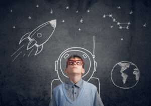 Kid with glasses on looking up into space with astronaut helmet drawn over his head on a chalkboard behind him.